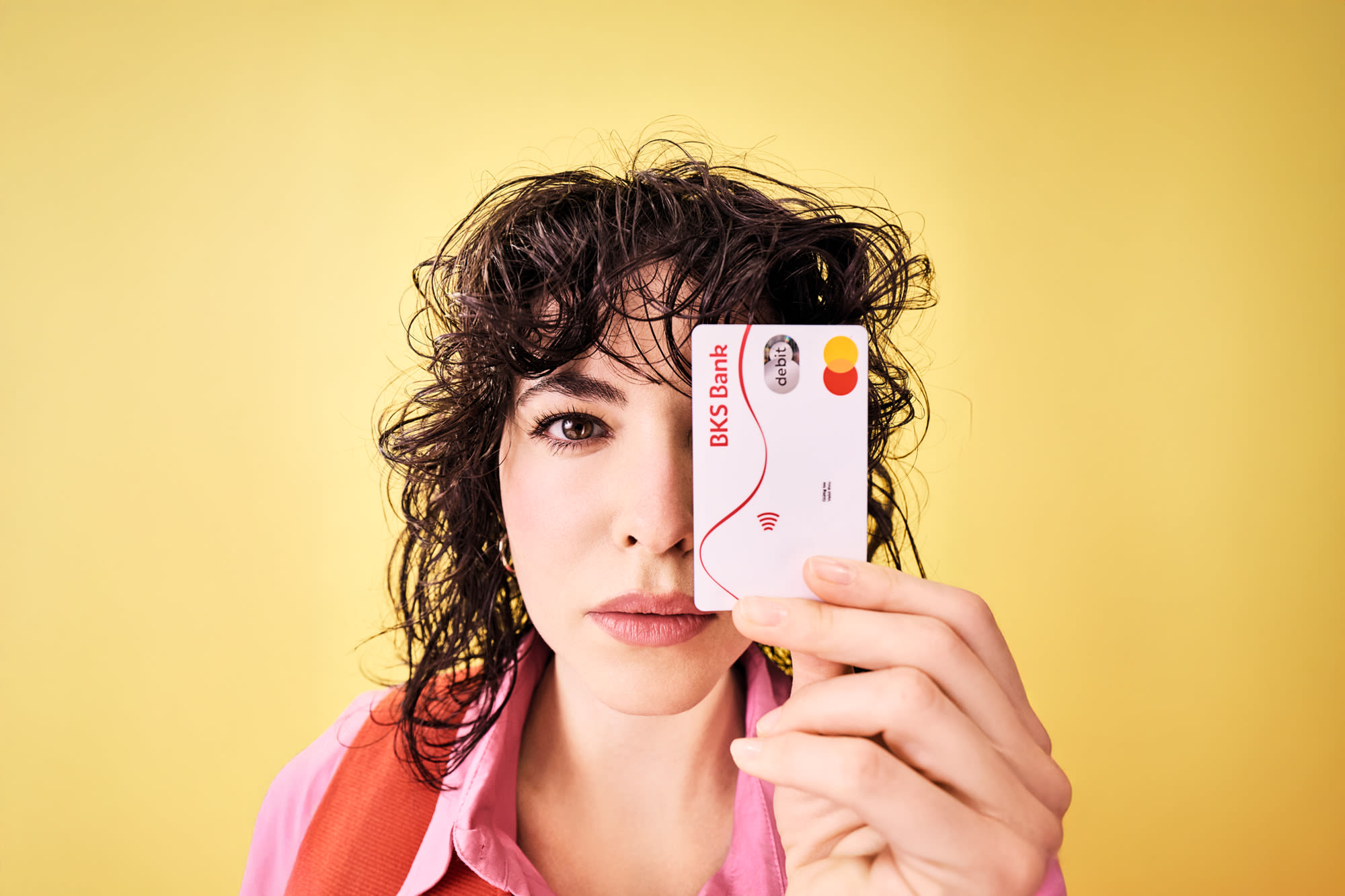 Žena drži BKS debitnu karticu ispred svog lica i stoji ispred žute pozadine. Žena drži BKS debitnu karticu ispred svog lica i stoji ispred žute pozadine.