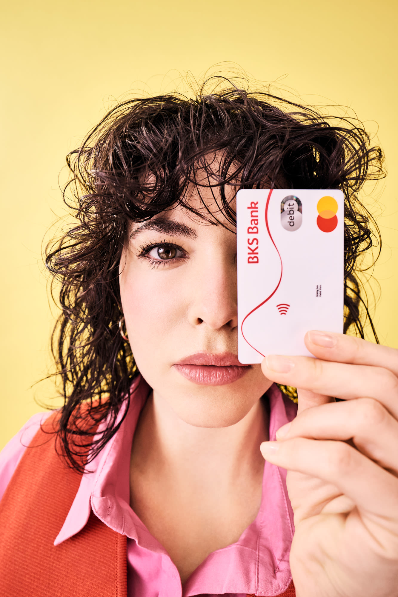 Žena drži BKS debitnu karticu ispred svog lica i stoji ispred žute pozadine. Žena drži BKS debitnu karticu ispred svog lica i stoji ispred žute pozadine.