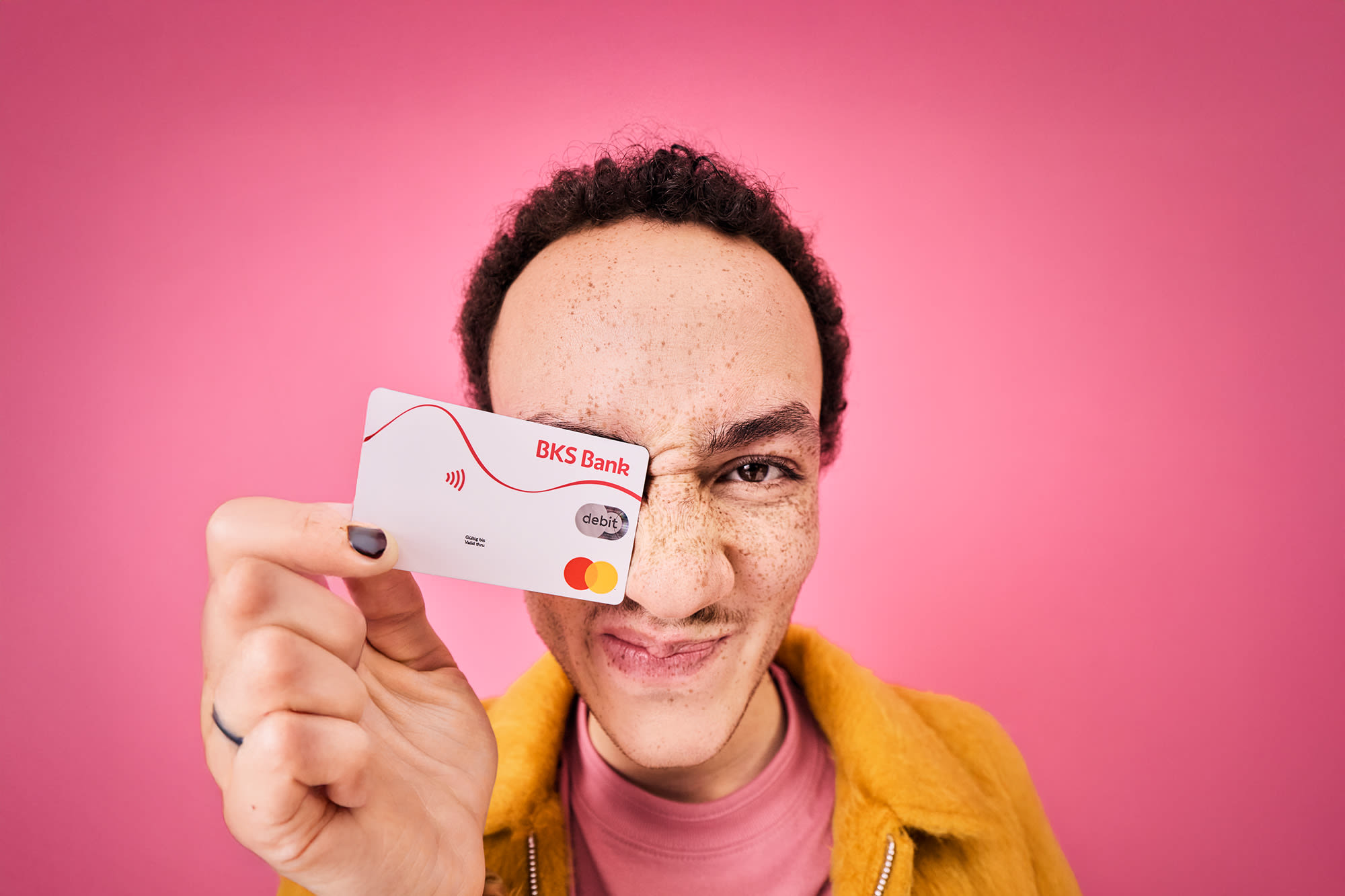 Muškarac drži BKS debitnu karticu ispred ružičaste pozadine – beskontaktno plaćanje s BKS Bankom. Muškarac drži BKS debitnu karticu ispred ružičaste pozadine – beskontaktno plaćanje s BKS Bankom.